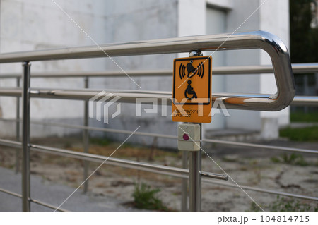 Yellow sign with a wheelchair sign and braille text with a button to call for help for enter on the railings in front of the ramp, accessible location 104814715
