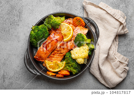 Healthy baked fish salmon steaks, broccoli, cauliflower, carrot in black cast iron casserole bowl on grey rustic stone background. Cooking a delicious low carb dinner, healthy nutrition concept Healthy baked fish salmon steaks, broccoli, cauliflower, carrot in black cast iron casserole bowl on grey rustic stone background. Cooking a delicious low carb dinner, healthy nutrition concept 104817817