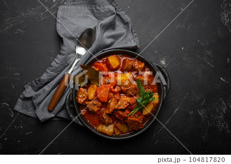 Beef meat stew with potatoes, carrot and delicious gravy in black casserole pot with bay leaves with spoon on black dark rustic concrete background from above . Beef meat stew with potatoes, carrot and delicious gravy in black casserole pot with bay leaves with spoon on black dark rustic concrete background from above . 104817820