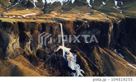 Aerial view of foss a sidu river and waterfall, water stream flowing off frozen hills and fields. Icelandic cascade in iceland running down off cliffs, scandinavian natural scenery. Slow motion. Aerial view of foss a sidu river and waterfall, water stream flowing off frozen hills and fields. Icelandic cascade in iceland running down off cliffs, scandinavian natural scenery. Slow motion. 104819255