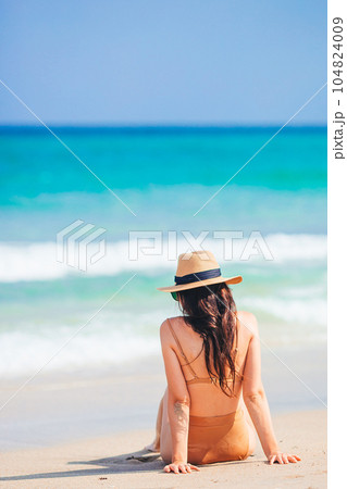 Young beautiful woman in swimsuit and straw hat on the beach vacation sitting on the sand 104824009