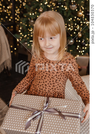 A little blonde girl is sitting on a soft bed in a festive interior decorated in a New Year's style. A child holds a Christmas gift in a craft package. A little blonde girl is sitting on a soft bed in a festive interior decorated in a New Year's style. A child holds a Christmas gift in a craft package. 104830370