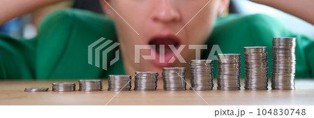 Shocked and surprised woman looks at stacks of coins on table. Shocked and surprised woman looks at stacks of coins on table. 104830748