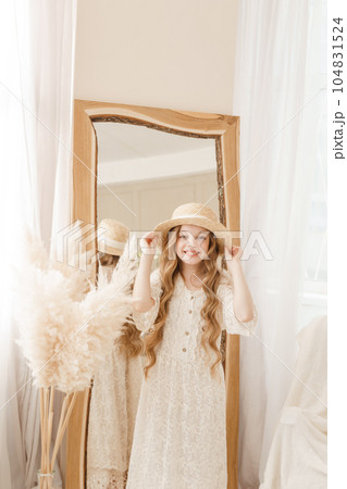 A beautiful teenage girl with long hair measures a straw hat in front of a mirror. Self-admiration of a blonde. A beautiful teenage girl with long hair measures a straw hat in front of a mirror. Self-admiration of a blonde. 104831524
