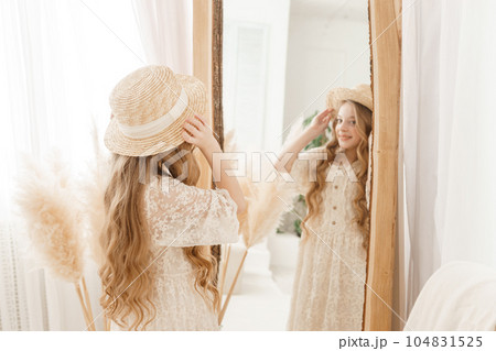 A beautiful teenage girl with long hair measures a straw hat in front of a mirror. Self-admiration of a blonde. 104831525