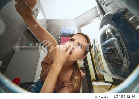 Young woman doing domestic chores, putting dirty clothes into washer. Bad smell. View from inside of washing machine Young woman doing domestic chores, putting dirty clothes into washer. Bad smell. View from inside of washing machine 104844244