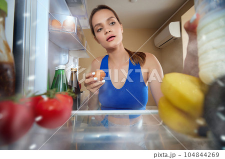 Breakfast time. Young woman opening fridge, taking egg and milk to prepare omelette for breakfast. View from inside of fridge Breakfast time. Young woman opening fridge, taking egg and milk to prepare omelette for breakfast. View from inside of fridge 104844269
