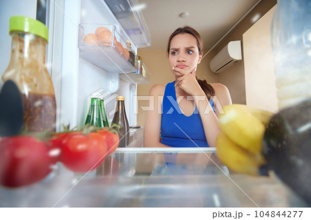 Young woman opening fridge, looking inside and choosing what to eat. Fridge filled with vegetables. View from inside of fridge Young woman opening fridge, looking inside and choosing what to eat. Fridge filled with vegetables. View from inside of fridge 104844277