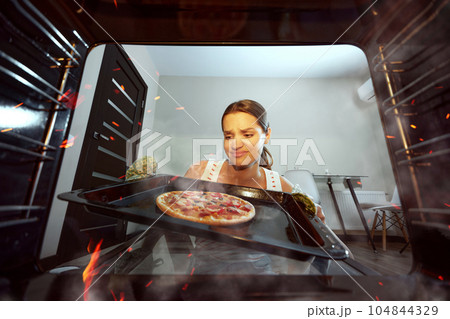 Young woman cooking, taking off pizza. Baking at home. Feeling bad about dish. Unpleasant look. View from oven Young woman cooking, taking off pizza. Baking at home. Feeling bad about dish. Unpleasant look. View from oven 104844329