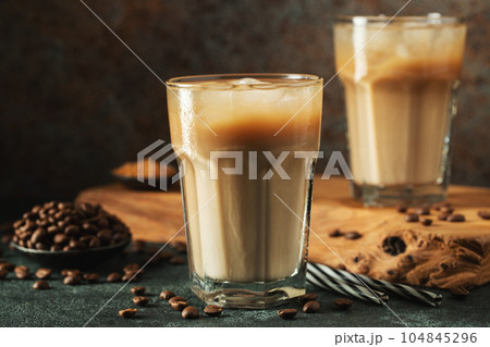 Ice coffee in a tall glass with cream poured over, ice cubes and beans on a dark concrete table. 104845296