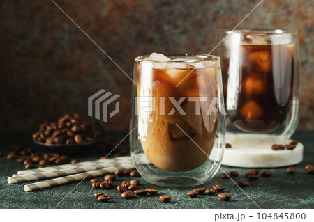 Ice coffee in a tall glass with cream poured over, ice cubes and beans on a dark concrete table. Ice coffee in a tall glass with cream poured over, ice cubes and beans on a dark concrete table. 104845800