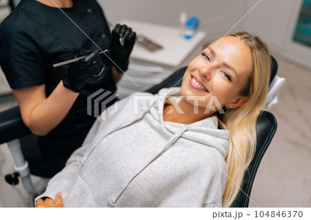 Portrait of charming young woman patient with perfect white teeth and smile satisfied after dental treatment lying on dental chair in dentistry clinic. Female dentist in gloves examining teeth. 104846370