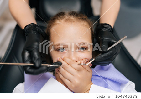 Close-up portrait of little cute child girl closing mouth by fear in dental clinic, frightened by dental drill, afraid of treatment. Cropped shot of unrecognizable female doctor holding dental tool. 104846388