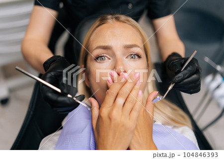 Closeup portrait of scared blonde young woman closing mouth by fear in dental clinic, afraid of treatment. Cropped shot of unrecognizable female doctor holding dental tool. Concept of dental treatment Closeup portrait of scared blonde young woman closing mouth by fear in dental clinic, afraid of treatment. Cropped shot of unrecognizable female doctor holding dental tool. Concept of dental treatment 104846393