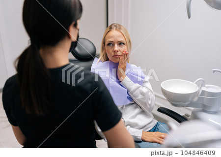 Back view of unrecognizable female dentist talking with sad young woman patient complaining of toothache holding hand on cheek at dentist office. Concept of stomatology, dentistry and orthodontics. 104846409
