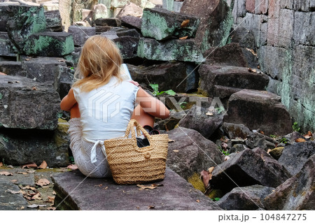 A young blonde European girl is sitting on a stone among medieval ruins and reading a book. 104847275