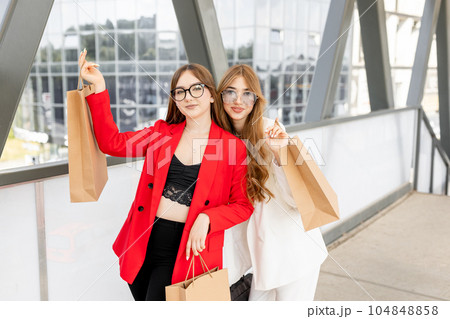Portrait of a two happy women dress in stylish suits are standing together in front of the shopping mall with a lots of paper bags. Concept of shopping Portrait of a two happy women dress in stylish suits are standing together in front of the shopping mall with a lots of paper bags. Concept of shopping 104848858