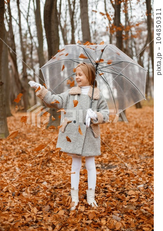 Little girl in fashion clothes standing in autumn forest. Girl posing for a photo and holding a transparent umbrella. Girl wearing grey coat and a headband. Little girl in fashion clothes standing in autumn forest. Girl posing for a photo and holding a transparent umbrella. Girl wearing grey coat and a headband. 104850311