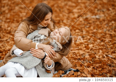 Young woman and little girl in autumn forest. Woman and her daughter sitting on a fallen leaves and hugging. Girl wearing fashion grey jacket and woman brown one. 104850315