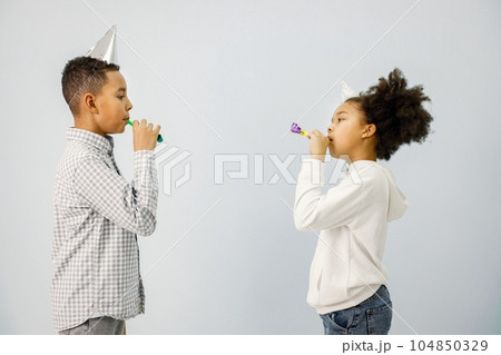 Boy and girl standing isolated on white background. Children blownig party horns. Black boy and girl wearing birthday cone hat. 104850329