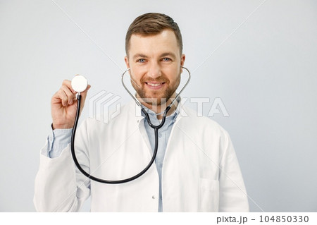 Male doctor standing isolated on white background. Portrait of a male physician looking at camera and smiling. Man wearing white medical gown and has stethoscope. 104850330