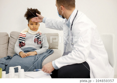 Male doctor in white medical gown asking sick girl about her symptoms. Black girl has fever and overspread blanket. Different medicines are on table near sofa. Male doctor in white medical gown asking sick girl about her symptoms. Black girl has fever and overspread blanket. Different medicines are on table near sofa. 104850357
