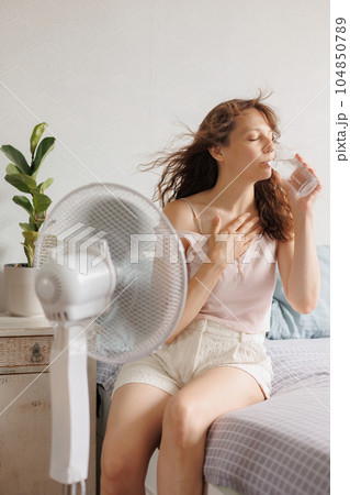 young European woman cools down at home with help of an air conditioner fan and drinks cool water  104850789