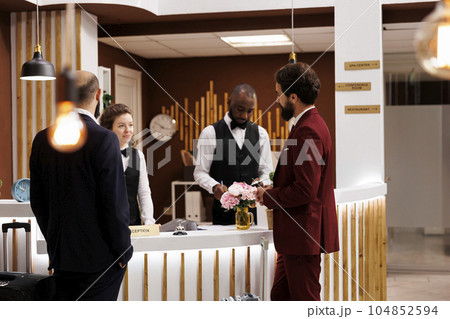 Professionals greeted by hotel staff at front desk, businessmen in suits arriving at luxury resort. People travelling for work partnership, doing check in procedure at reception. 104852594