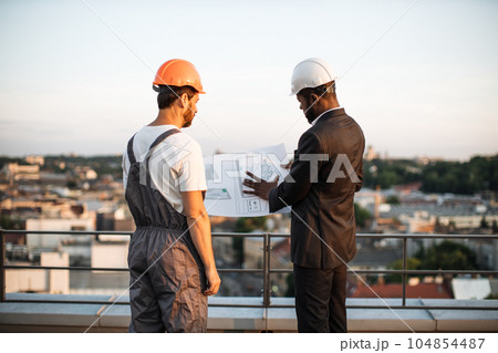 Workman presenting blueprints to urban planner on rooftop 104854487