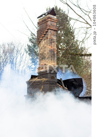 Remains of a house after a fire. The brick tube of the stove is shrouded in dense smoke. A vertical view of the gloomy ash Remains of a house after a fire. The brick tube of the stove is shrouded in dense smoke. A vertical view of the gloomy ash 104863608