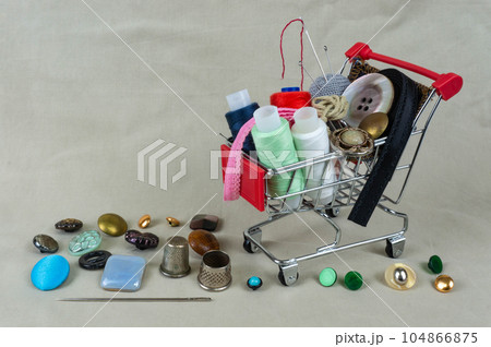 A shop basket with threads, sewing accessories and buttons. Next to thimbles and tools for sewing and embroidery. The background is made of gray-beige fabric. Place for text or logo. 104866875