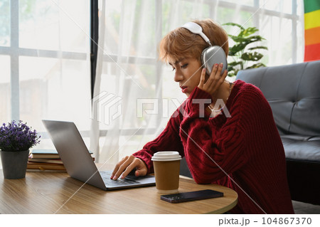 Smiling asian teenage gay man wearing headphone watching movies or studying online on laptop at home 104867370