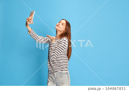 Portrait of young beautiful girl in casual clothes taking selfie with mobile phone against blue studio background. Social media 104867851