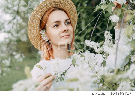 Portrait of a woman in a straw hat in a cherry blossom. Free outdoor recreation, spring blooming garden. 104868717