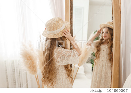 A beautiful teenage girl with long hair measures a straw hat in front of a mirror. Self-admiration of a blonde. 104869007