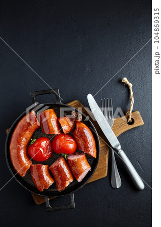 sliced fried pork sausage in a cast iron pan with tomatoes and cutlery on a wooden cutting board on a dark concrete table 104869315