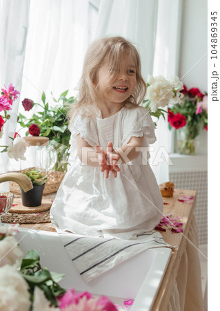 A little blonde girl on a kitchen countertop decorated with peonies. Spring atmosphere. A little blonde girl on a kitchen countertop decorated with peonies. Spring atmosphere. 104869345