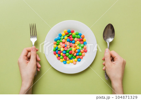 the girl holds cutlery in her hands and eats sweets in a plate. Health and obesity concept, top view on colored background the girl holds cutlery in her hands and eats sweets in a plate. Health and obesity concept, top view on colored background 104871329