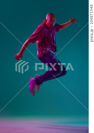Full-length portrait of young bald man with moustache in stylish clothes emotionally jumping against cyan studio background in neon light Full-length portrait of young bald man with moustache in stylish clothes emotionally jumping against cyan studio background in neon light 104871548