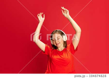 Portrait of young beautiful girl in casual clothes listening to music in headphones and dancing against red studio background 104874828
