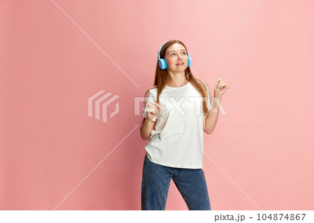 Portrait of young beautiful girl in casual clothes listening to music in headphones against pink studio background Portrait of young beautiful girl in casual clothes listening to music in headphones against pink studio background 104874867