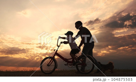 happy family in the park. father teaching son to ride a bike at sunset silhouette in the park. son child learning to ride a bike at sunset father helping son. child playing riding a lifestyle bike 104875503