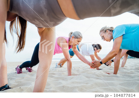 Women of various ages doing fitness workouts in class exercise with coach on beach. Ladies doing paired plank exercises and high-fiving each other. Sport for health and wellbeing. Active lifestyle Women of various ages doing fitness workouts in class exercise with coach on beach. Ladies doing paired plank exercises and high-fiving each other. Sport for health and wellbeing. Active lifestyle 104877814