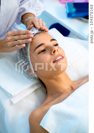 Cosmetologist preparing client for cosmetic procedures by putting disposable cap on woman head 104878168