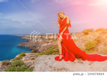 woman red silk dress stands by the ocean, with mountains in the background, as her dress sways in the breeze. 104878664