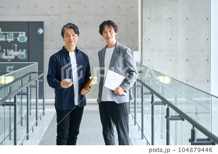 オフィスの通路でノートパソコンを持って立つ2人のビジネスパーソン オフィスの通路でノートパソコンを持って立つ2人のビジネスパーソン 104879446