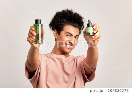 Portrait of young handsome smiling guy with bottles of moisturizing facial lotion and healing creme in his hands. Swarthy man with black curly hair uses modern cosmetics in his skincare routine. 104880278