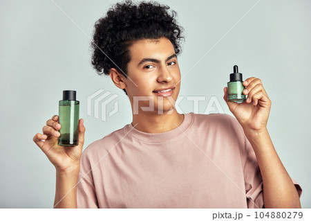 Portrait of young handsome smiling guy with bottles of moisturizing facial lotion and healing creme in his hands. Swarthy man with black curly hair uses modern cosmetics in his skincare routine. 104880279