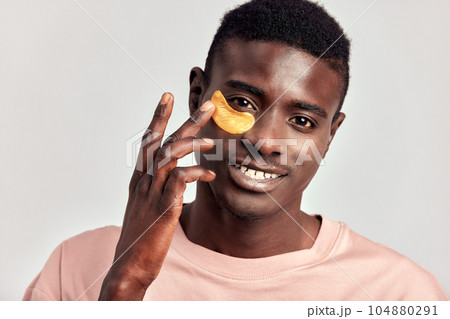 Young handsome black guy applying cosmetic under eye hydrogel patch on his face. African American millennial man practices skin care routine to keep healthy and youthful looking. Studio portrait. Young handsome black guy applying cosmetic under eye hydrogel patch on his face. African American millennial man practices skin care routine to keep healthy and youthful looking. Studio portrait. 104880291