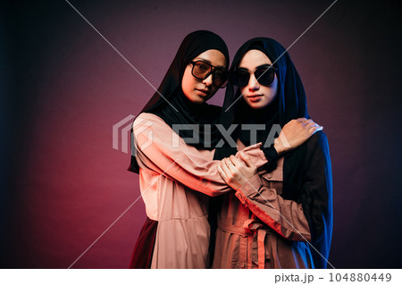 two muslim women wearing hijab dress and sunglasses hugging looking at camera two muslim women wearing hijab dress and sunglasses hugging looking at camera 104880449